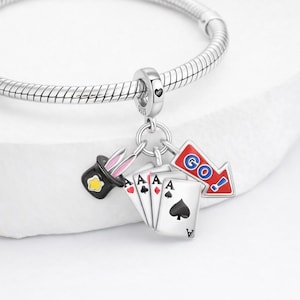 Puede incluir: Pulsera de plata con una colección de abalorios. Los abalorios incluyen un sombrero de copa negro con orejas de conejo rosas, naipes con símbolos de as y una flecha roja con la palabra "GO!" en azul.