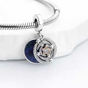 May include: A silver charm with a celestial design. The charm features a dark blue enamel crescent moon, a circle of clear crystals, and a rose gold star in the centre. Several smaller stars are also present. The charm is attached to a silver bracelet.