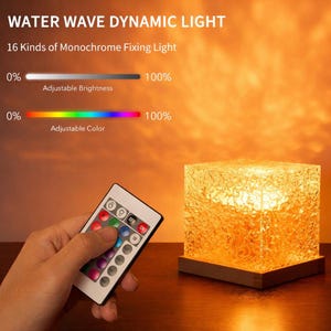 Puede incluir: Una lámpara cúbica con superficie texturizada, que emite un brillo naranja cálido. La lámpara se encuentra sobre una base de madera. Se sostiene un mando a distancia cerca, con botones para ajustar el brillo y el color. El texto de la imagen dice "WATER WAVE DYNAMIC LIGHT".