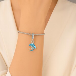Könnte beinhalten: Ein silbernes Charm-Armband mit einem blau-weißen Eidechsen-Charm. Der Eidechsen-Charm hat ein geometrisches Design und ist mit kleinen Kristallen verziert. Das Armband wird an einem Arm gezeigt, mit einem weißen Oberteil im Hintergrund.