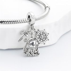 May include: A silver charm with Viking-themed pendants. The charm features a Viking ship, an axe, and a ship's wheel with engraved text. The charm is attached to a silver bracelet.