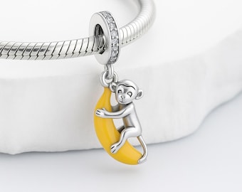 Puede incluir: Un charm de plata con un mono abrazando un plátano amarillo. El charm está unido a una pulsera de plata. El anillo del charm está incrustado con pequeñas piedras transparentes.