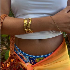 Peut inclure: Un bracelet manchette en or, de forme ondulée et abstraite. Le bracelet est porté au poignet d'une personne portant un débardeur blanc et un sarong coloré.