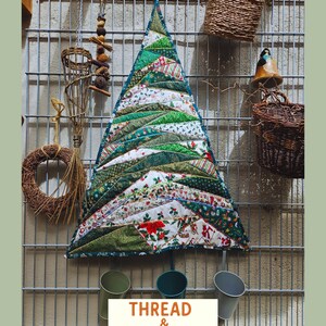 Peut inclure: Décoration murale en forme de sapin de Noël, faite de tissus verts et blancs à motifs variés. Le sapin est accroché sur une grille métallique avec d'autres éléments décoratifs. Le texte "THREAD & MEMORY" est affiché en bas.