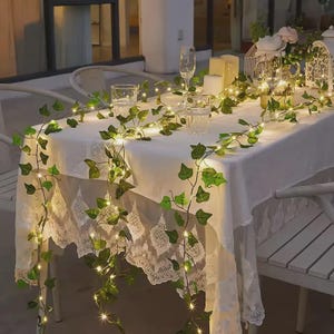 Può includere: Un tavolo apparecchiato per un evento all'aperto, drappeggiato con una tovaglia di pizzo bianco. Ghirlande di edera verde con piccole luci bianche calde sono drappeggiate sul tavolo. Sono presenti anche bicchieri e candele trasparenti.