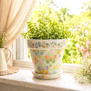 May include: A tapered ceramic plant pot with a matching saucer, featuring a repeating pattern of colorful butterflies in orange, yellow, blue, and green against a light background, topped with a decorative floral border around the rim.