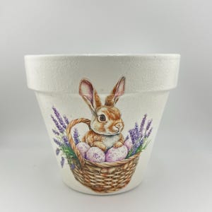May include: A white ceramic flower pot with a textured finish. The pot features a detailed illustration of a brown bunny sitting in a woven basket filled with purple speckled eggs. Lavender sprigs frame the basket.
