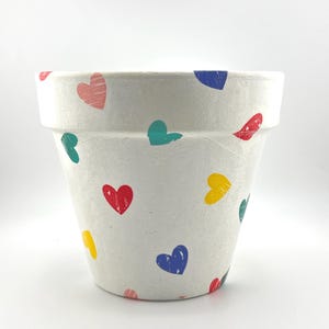 May include: White ceramic flower pot decorated with colorful heart shapes in red, pink, blue, yellow, and teal. The pot has a wide rim and a slightly tapered body, suitable for planting flowers or herbs.