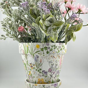 May include: A decorative flower pot with a matching saucer, filled with an assortment of artificial flowers and greenery. The pot is white with a colorful floral and Easter-themed pattern featuring bunnies, ducks, and butterflies.