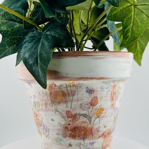 May include: A decorative flower pot with a distressed white finish, featuring a painted design of brown rabbits, flowers, and butterflies. The pot is filled with green ivy plants. The rim has a brown accent.
