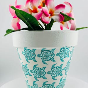 May include: White ceramic flower pot with a teal turtle pattern. The pot contains pink and white flowers with green leaves. The pot is approximately 6 inches tall and 6 inches wide.