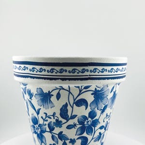 May include: A white and blue floral patterned ceramic flower pot. The pot features a detailed blue floral design on a white background. The rim has a blue border with a decorative pattern.