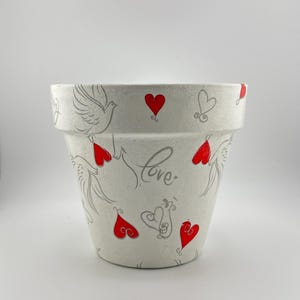 May include: White ceramic flower pot decorated with red hearts, gray doves, and the word "love". The pot has a wide rim and a slightly tapered shape, suitable for planting flowers or herbs.