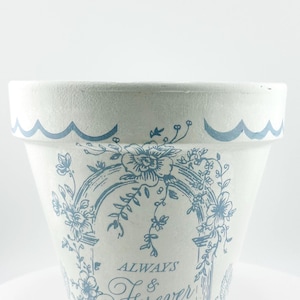May include: White ceramic flower pot with a blue floral design and the words "Always & Forever." The rim has a wavy blue border. The pot is suitable for indoor or outdoor use.