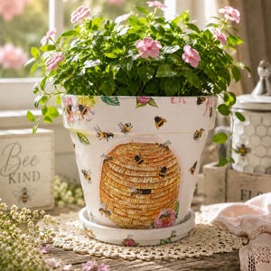 May include: A white ceramic flower pot featuring a painted golden beehive, multiple bees, and pink floral accents. The pot includes a matching saucer and contains a green plant with small pink flowers, sitting on a wooden surface.