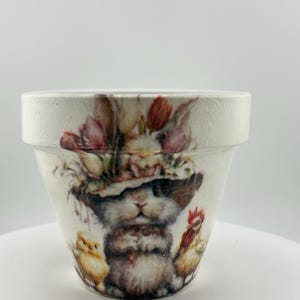 May include: A white ceramic flower pot with a detailed illustration of a bunny wearing a floral hat, with a chick and a rooster. The pot has a textured finish and a wide rim, perfect for spring decor.