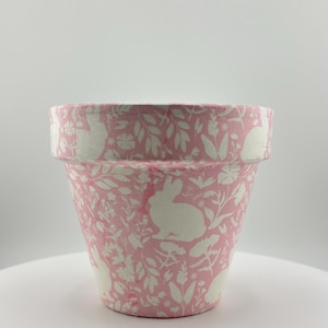 May include: A pink flower pot with a white rabbit and floral pattern. The pot is cylindrical with a slightly flared rim. The design features silhouettes of rabbits and various plants, creating a spring or Easter theme.