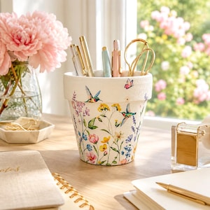 May include: A white ceramic pencil holder featuring a colorful floral pattern with hummingbirds and butterflies. The cylindrical container holds several pens and gold-toned scissors, standing on a wooden surface near a glass vase of pink flowers.