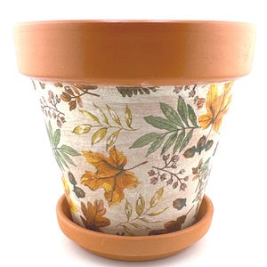 May include: A terracotta-colored flower pot with a matching saucer. The pot is decorated with a cream-colored background and a pattern of autumn leaves in shades of yellow, orange, and green. The pot has a wide rim.