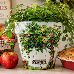 May include: A white ceramic plant pot featuring a colorful illustration of an apple tree with a wooden swing, two cats, and a basket of apples. A lush green plant grows from the top of the pot, which is decorated with white blossoms and red fruit.