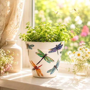 May include: A white ceramic flower pot featuring colorful illustrations of dragonflies in shades of blue, green, purple, and orange. The pot contains a small green plant and sits on a surface, showcasing detailed dragonfly artwork.
