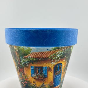 May include: A decorative terracotta flower pot with a vibrant scene of a yellow house with blue shutters and door, a bicycle, and lush greenery. The rim of the pot is painted a solid blue color. Ideal for indoor or outdoor use.