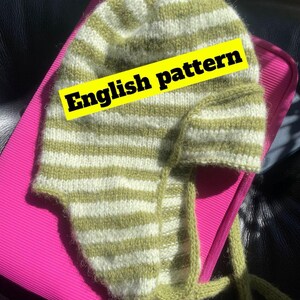 Könnte beinhalten: Eine gestrickte Mütze mit Ohrenklappen, mit horizontalen Streifen in Hellgrün und Weiß. Die Mütze hat lange Bänder und den Text "English pattern" auf einem gelben Banner.