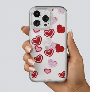 May include: Transparent phone case featuring a pattern of red, pink, and white crocheted hearts. The case is held in a hand against a light background. The hearts are of varying sizes and are spread across the case.