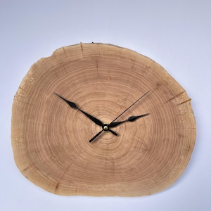 Könnte beinhalten: Eine Holzuhr mit schwarzen Zeigern und einem goldenen Zentrum. Das Zifferblatt der Uhr ist eine natürliche Holzscheibe mit sichtbaren Jahresringen.