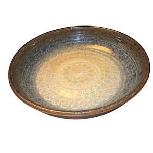 Puede incluir: Un cuenco de cerámica hecho a mano con un diseño ancho y poco profundo. El cuenco presenta un centro beige claro con un patrón circular detallado. El borde es de color marrón oscuro, que pasa a un tono azul grisáceo hacia el centro. El cuenco tiene aproximadamente 5 cm de profundidad.
