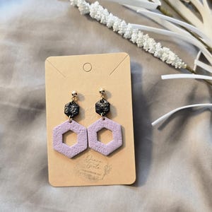 Könnte beinhalten: Handgefertigte Ohrringe mit geometrischem Design. Die Ohrringe haben eine schwarz strukturierte Oberseite und eine hellviolette Sechseckform. Die Ohrringe werden auf einer braunen Karte mit der Signatur des Künstlers präsentiert.