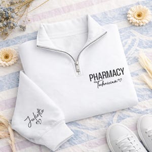 May include: A white quarter-zip sweatshirt featuring black text on the chest that reads PHARMACY Technician with a small heart icon. The sleeve cuff displays the name Juliette in black script lettering followed by a small heart symbol.