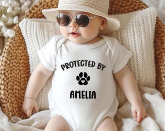 May include: A baby wearing a white onesie with the text "PROTECTED BY" above a paw print and heart design, and the name "AMELIA" below. The baby is wearing a straw hat and sunglasses, and is sitting in a woven basket.