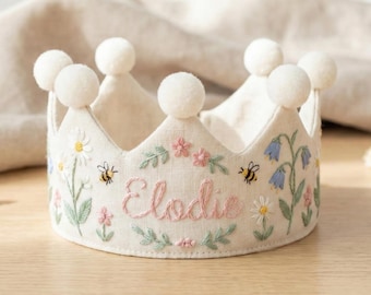 May include: A cream-colored fabric crown with embroidered floral details, including daisies, pink flowers, and bluebells. The name "Elodie" is embroidered in pink script. The crown is topped with white pom-poms.