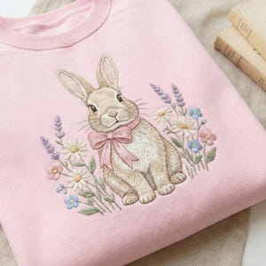 May include: A light pink sweatshirt featuring an embroidered bunny wearing a pink bow, surrounded by embroidered flowers. The bunny is light brown with long ears. The flowers include daisies, lavender, and other colorful blooms.