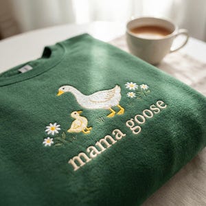 May include: A dark green sweatshirt with an embroidered design featuring a white goose, a yellow gosling, and the words "mama goose" in cream-colored thread. Small white daisies are also embroidered around the goose and gosling.