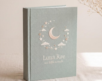 Puede incluir: Un libro de recuerdos para bebés de color verde claro con las palabras "Luna Rae" y "our little miracle" impresas en la portada. La portada presenta un diseño bordado de una luna creciente, estrellas y nubes. El libro está de pie.