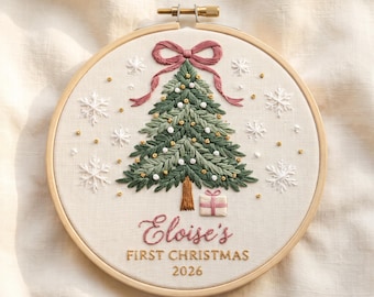 Puede incluir: Un bastidor de bordado redondo de madera que contiene una pieza de tela con un árbol de Navidad verde bordado a mano, un lazo rosa, copos de nieve blancos y un pequeño regalo. El texto dice Eloise's FIRST CHRISTMAS 2026 en hilo rosa y dorado.