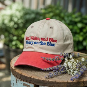 May include: A beige baseball cap with a red brim and button. The cap features the embroidered text "Red, White, and Blue Heavy on the Blue" in red and blue. The cap is resting on a wooden surface with flowers.