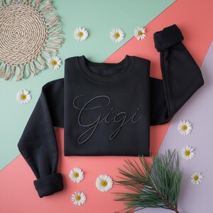 K&ouml;nnte beinhalten: Schwarzes Sweatshirt mit dem Namen "Gigi" in Schreibschrift gestickt. Das Sweatshirt wird auf einem farbenfrohen Hintergrund mit G&auml;nsebl&uuml;mchen und einer gewebten Matte pr&auml;sentiert.