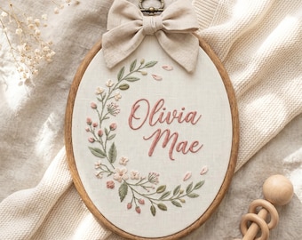 Puede incluir: Aro de bordado ovalado con el nombre "Olivia Mae" en escritura rosa, rodeado de bordados florales en rosa y verde. Un lazo beige está adjunto en la parte superior, y el aro tiene un marco de madera. Un sonajero de madera está en el fondo.