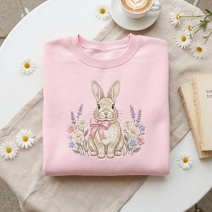 May include: A light pink sweatshirt featuring an embroidered bunny wearing a pink bow, surrounded by flowers. The bunny is light brown and white. The flowers include daisies, lavender, and other colorful blooms. The sweatshirt is folded on a table.