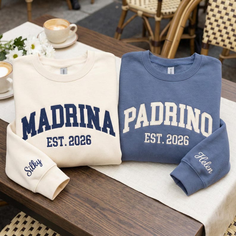 May include: Two crewneck sweatshirts, one cream and one blue, with embroidered text. The cream sweatshirt says "Madrina" and the blue one says "Padrino", both with "EST. 2026". The cream sleeve says "Silky" and the blue sleeve says "Helen".