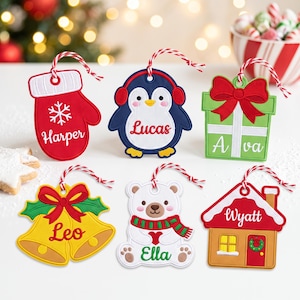 May include: Six embroidered Christmas ornaments with red and white striped hanging strings. Designs include a red mitten labeled Harper, a penguin labeled Lucas, a green gift box labeled Ava, yellow bells labeled Leo, a bear labeled Ella, and a house labeled Wyatt.