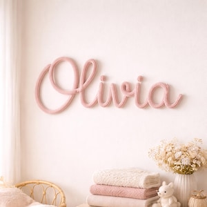以下が含まれることがあります： ピンク色のニットの壁掛けで、名前「Olivia」を筆記体で表現。白い壁に設置され、毛布の山と花瓶の上に配置されています。テーマは子供部屋の装飾です。