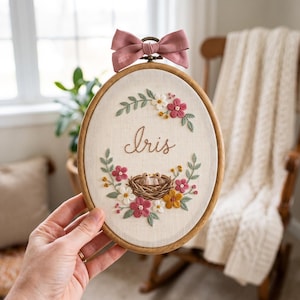 May include: An oval wooden embroidery hoop containing white fabric with the name Iris stitched in brown thread. A floral wreath surrounds the name and a small bird nest with two birds. A dusty rose fabric bow is attached to the top of the hoop.