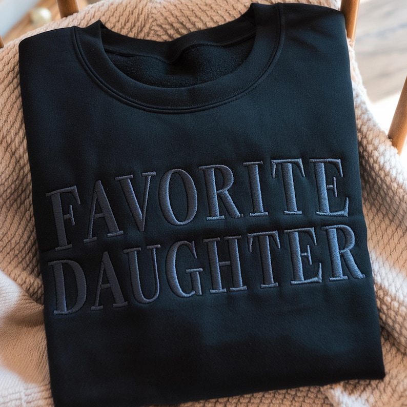 K&ouml;nnte beinhalten: Ein schwarzes Sweatshirt mit dem gestickten Text "FAVORITE DAUGHTER" in einem dunkleren Grauton. Das Sweatshirt liegt gefaltet auf einer hellfarbigen Decke, die auf einem Holzstuhl liegt.