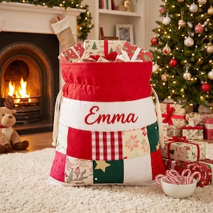 May include: A large fabric gift sack with a drawstring closure, featuring a red top section and a patchwork lower body with red, white, and green patterns. The name Emma is embroidered in red script on a white textured band across the center.