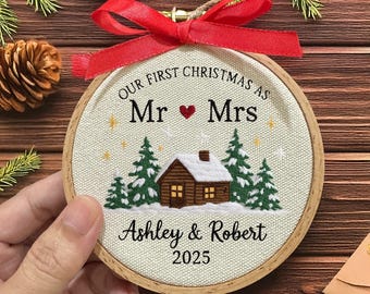 May include: A round, embroidered Christmas ornament with a wooden frame and a red ribbon. The ornament features the text "OUR FIRST CHRISTMAS AS Mr ❤️ Mrs" above a cabin scene and the names "Ashley & Robert 2025".