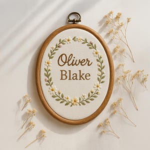 May include: Oval wooden embroidery hoop with the name "Oliver Blake" stitched in brown thread. The name is surrounded by a wreath of white daisies and green leaves. A small brass ring is at the top for hanging.